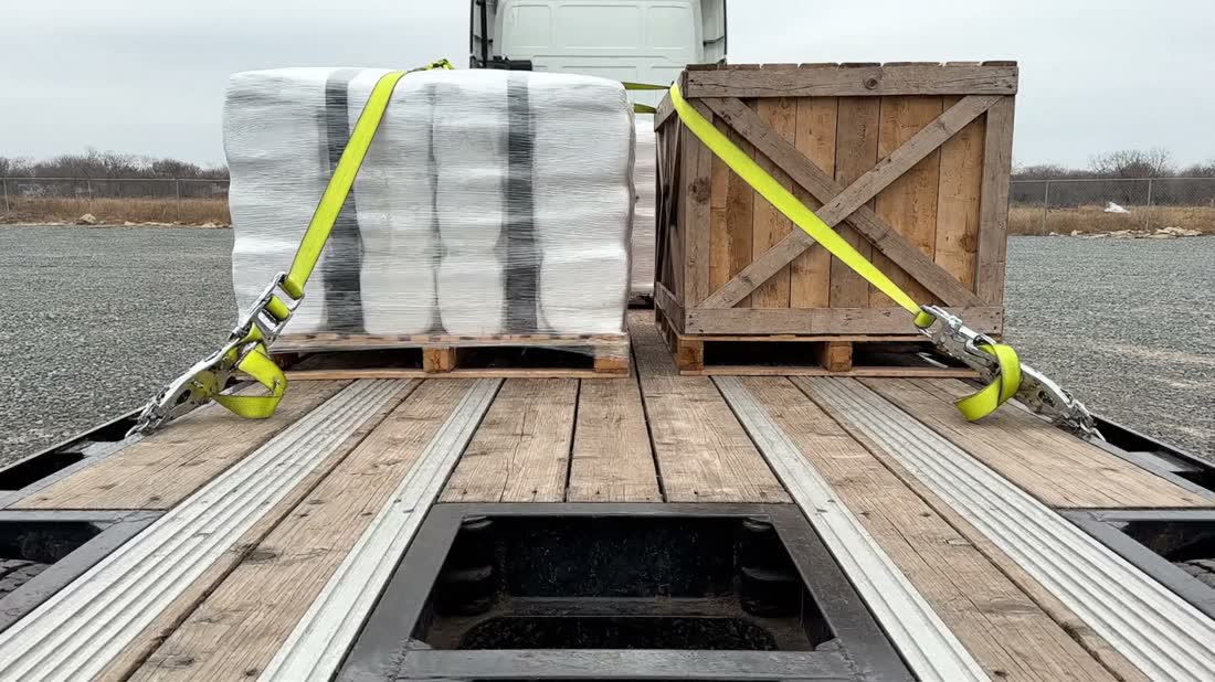 A shrink-wrapped pallet and a wooden crate sitting on a flatbed trailer deck, each pulled down by a yellow ratchet strap running to the side rail.